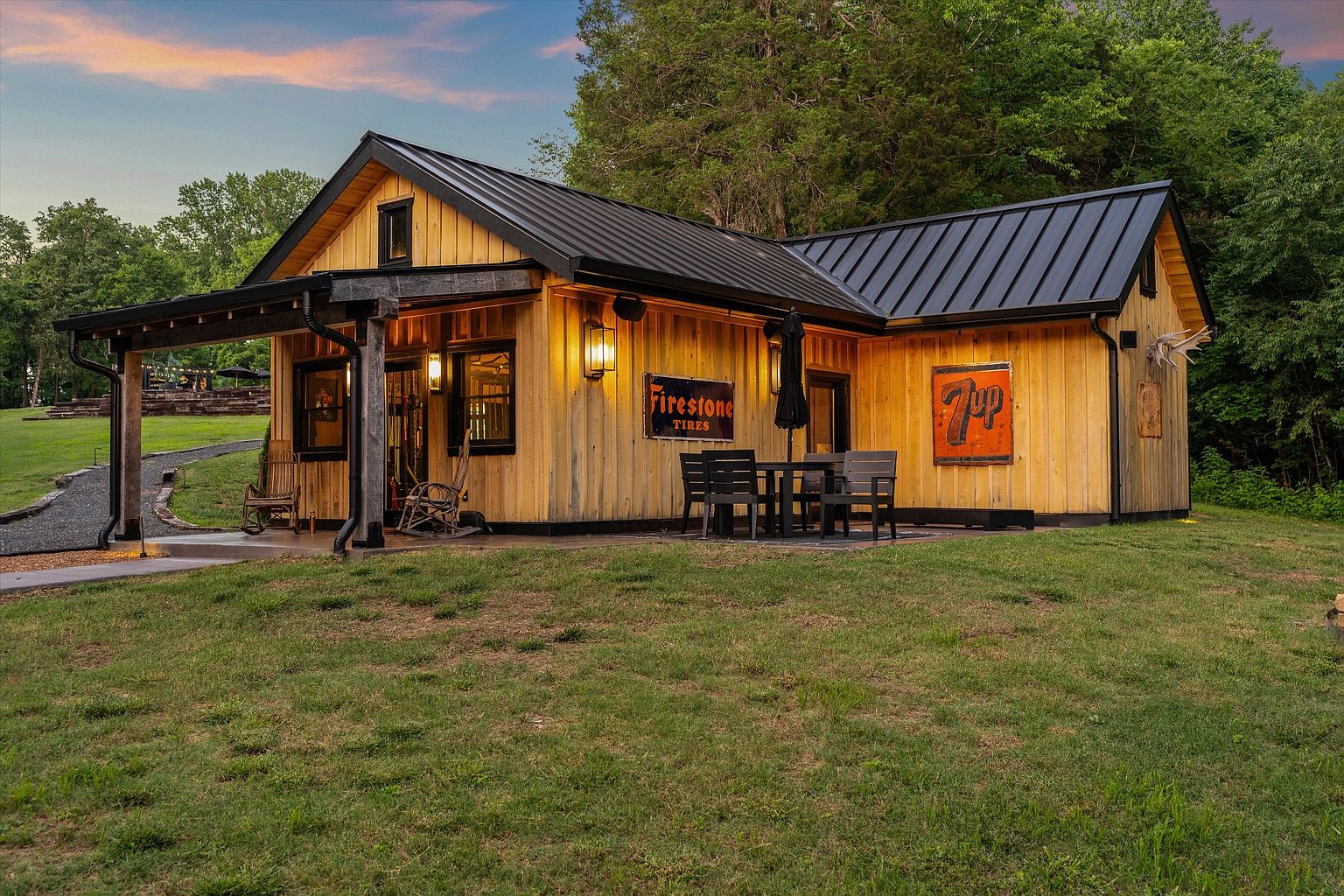 This is a charming front view of a rustic-style building with a black metal roof and natural wood siding. The exterior features vintage signage, including a 'Firestone Tires' sign, adding character. A covered porch with rocking chairs and an outdoor dining area enhance the inviting atmosphere, set against a backdrop of lush greenery.
