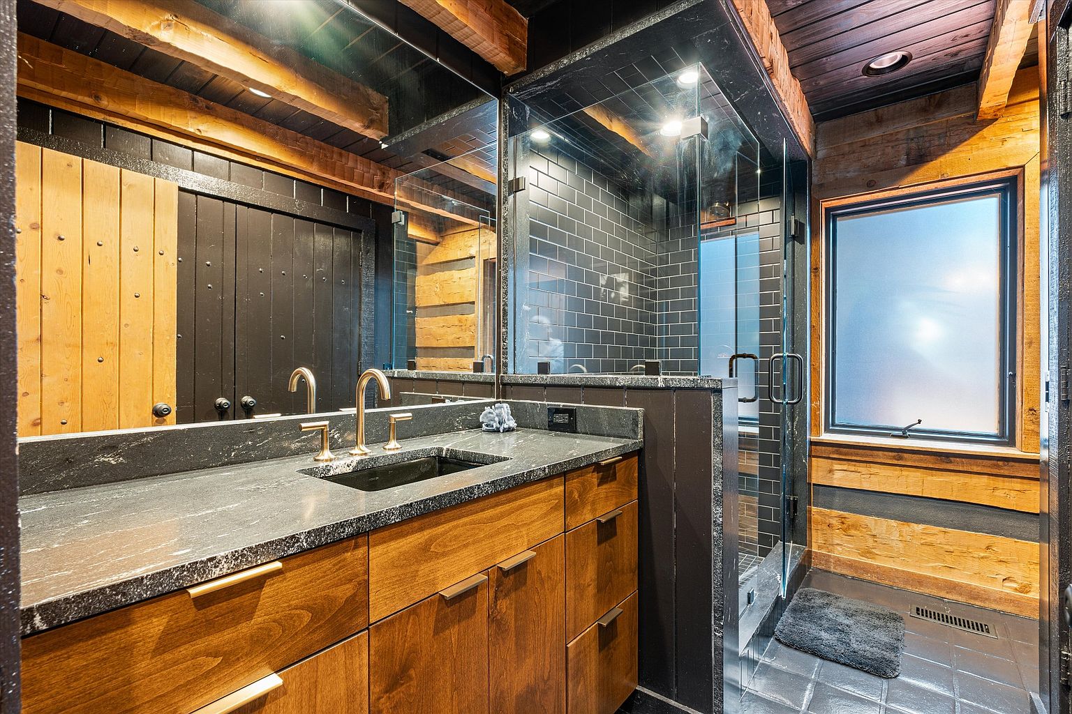 This is a primary bathroom featuring a double vanity with a dark countertop and wooden cabinets. A glass-enclosed shower with dark tiling is adjacent to the vanity. The bathroom has a rustic-modern style with exposed wooden beams and a frosted window providing natural light.