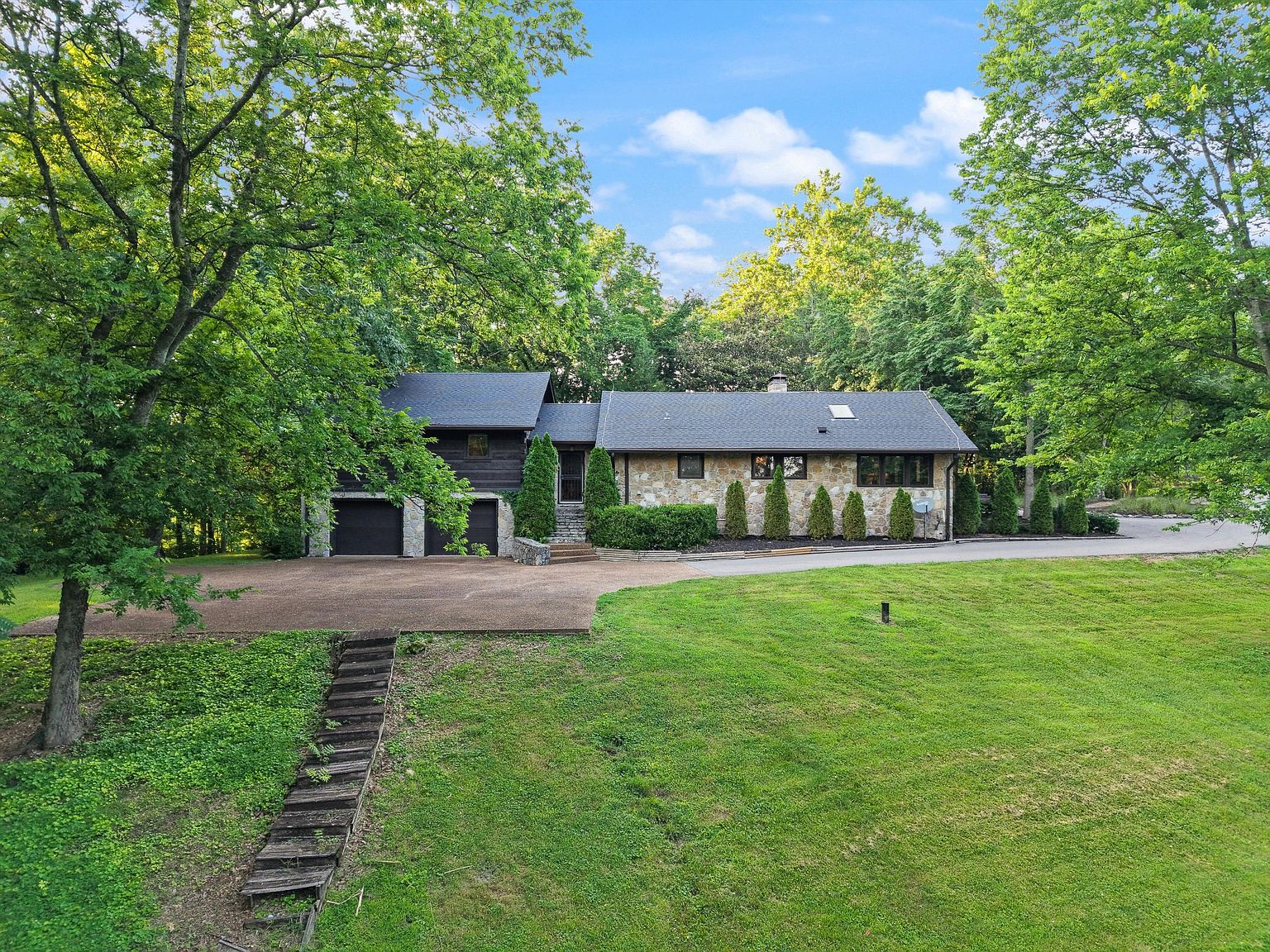 This is an exterior front view of a single-family home. The house features a combination of stone and dark wood siding, with a dark roof and a two-car garage on the left. The front yard is expansive and well-maintained, with mature trees framing the property, and a set of wooden stairs leading up to the driveway.