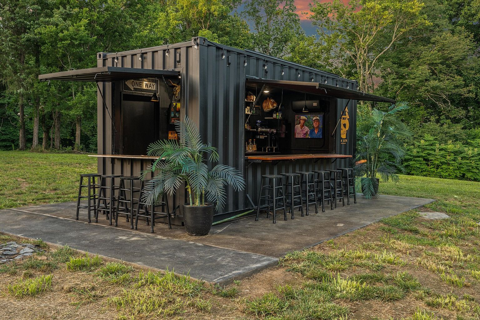 This image showcases a unique outdoor bar constructed from a shipping container, set in a lush yard. The bar features open serving windows with awnings, bar stools, and decorative plants, creating an inviting entertainment space. The surrounding greenery and concrete patio enhance the bar's appeal, making it a standout feature for outdoor gatherings.