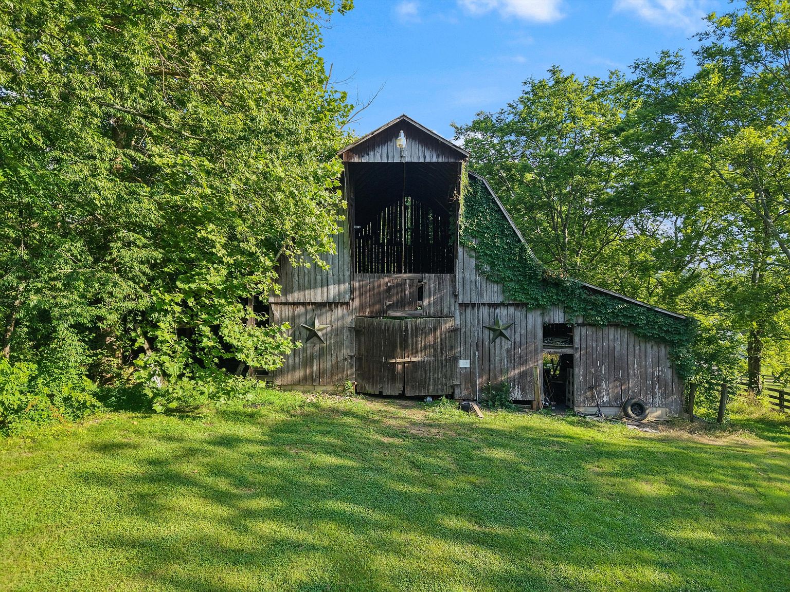The image showcases a rustic barn nestled amidst lush greenery, creating a charming and idyllic scene. The barn features weathered wood, a high roofline, and decorative star accents, adding to its character. The surrounding yard is covered in vibrant green grass, enhancing the property's appeal and suggesting a peaceful, rural setting.