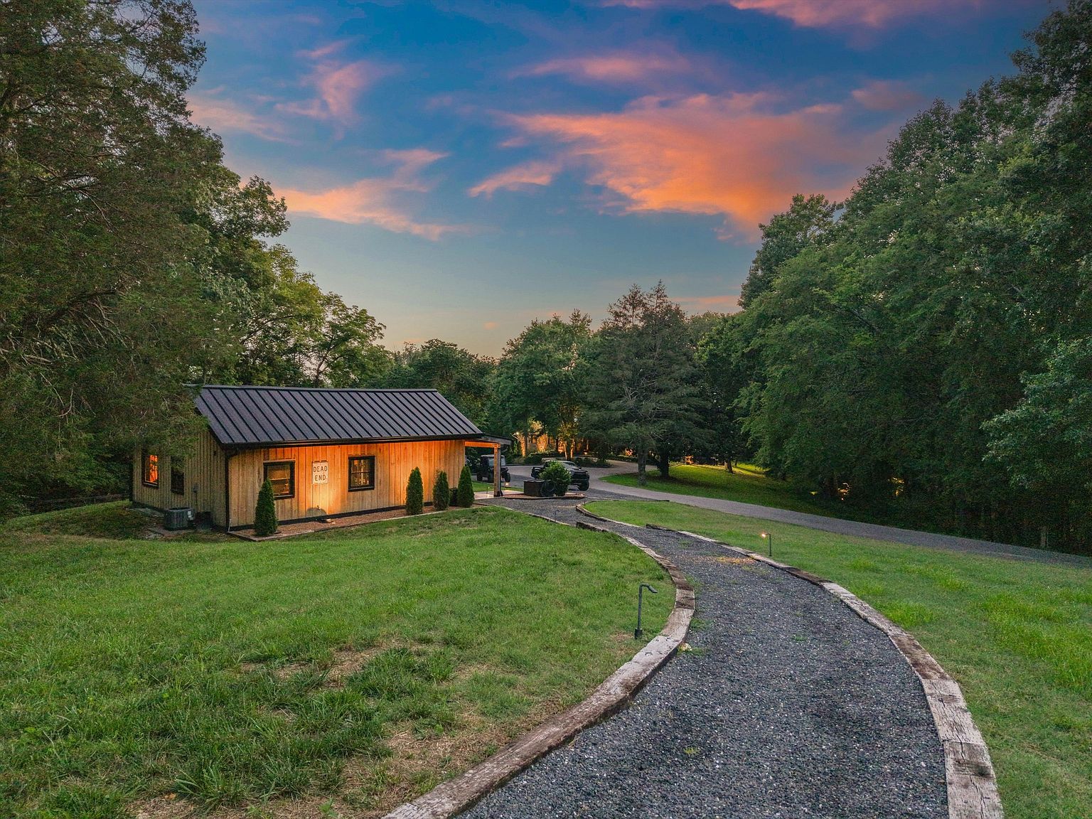 This image showcases the front exterior of a charming, modern cabin-style home. The house features natural wood siding, a dark metal roof, and a well-manicured lawn. A gravel pathway bordered by wooden planks leads to the house, enhancing its inviting appeal and highlighting the property's serene setting amidst lush greenery and a colorful sunset sky.