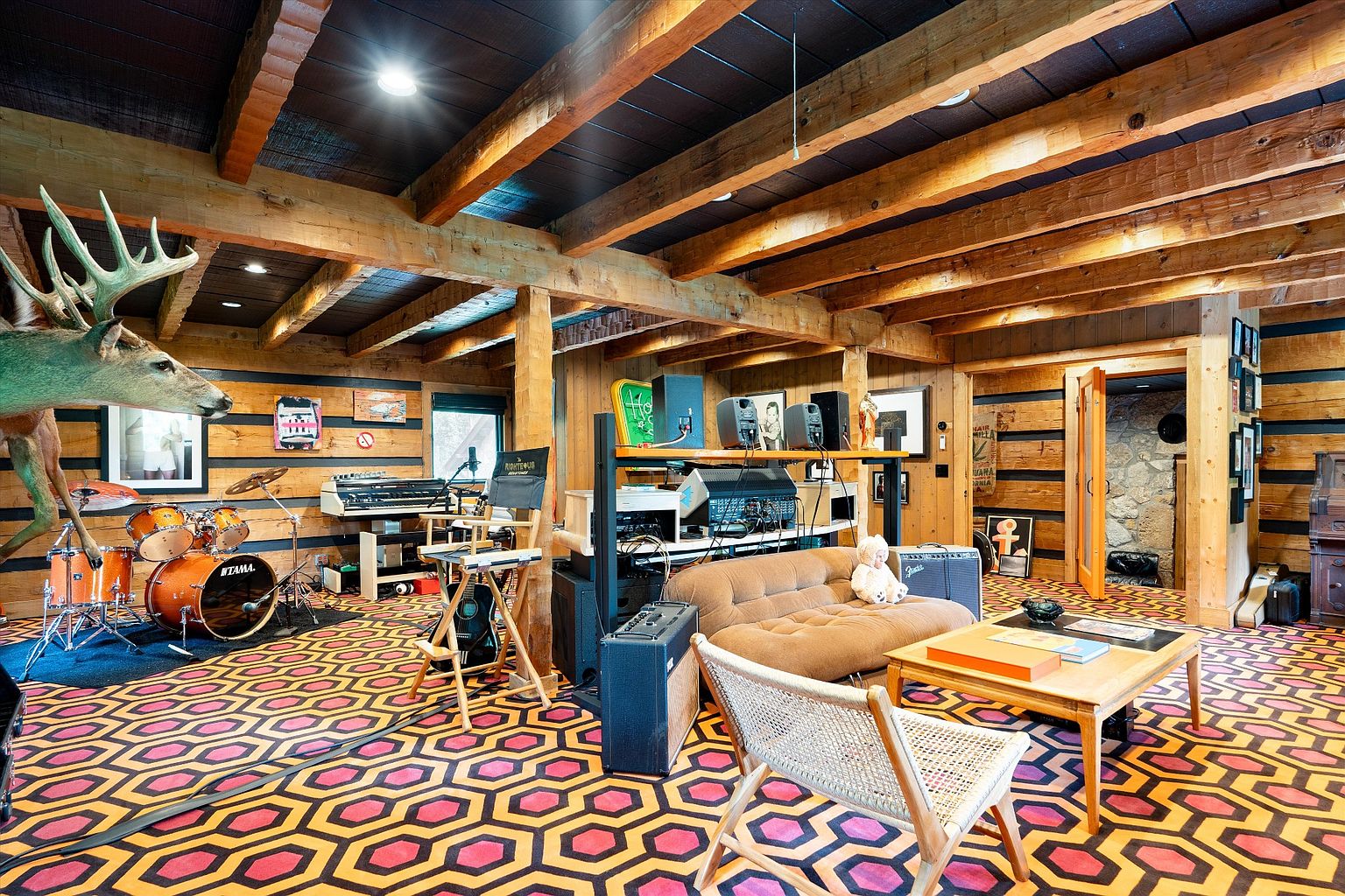 This unique living room features a striking geometric patterned floor, wood-paneled walls, and exposed wooden beams on the ceiling, creating a rustic yet artistic atmosphere. The room is furnished with a brown sofa, a wooden coffee table, and a drum set, suggesting a space for both relaxation and creative expression. A mounted deer head adds a distinctive touch to the room's decor.