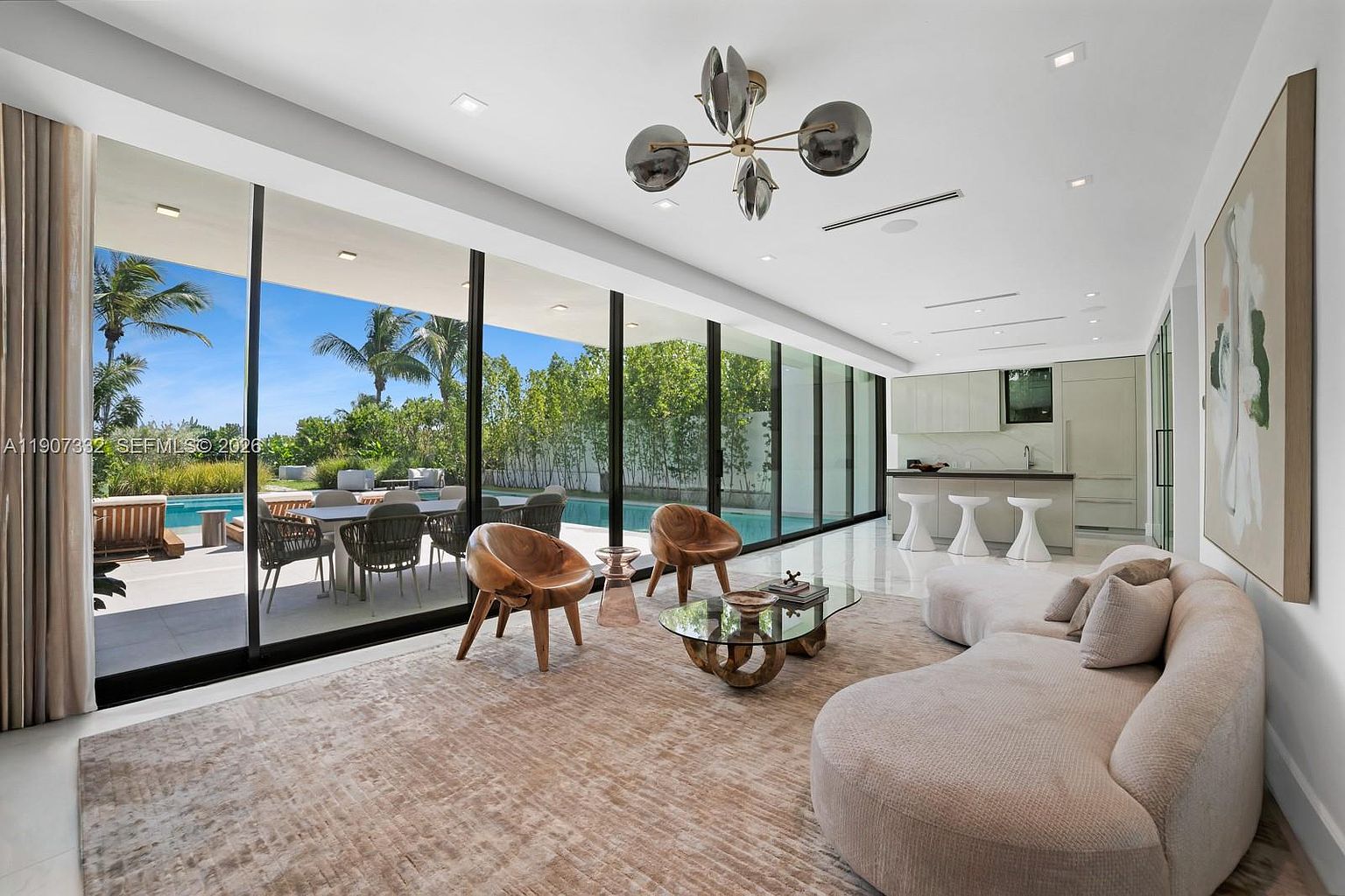 This bright, open-concept living area features a curved sofa, two sculptural wooden chairs, and a glass-topped coffee table set upon a textured area rug. The space is defined by expansive floor-to-ceiling glass walls that provide a seamless transition to the outdoor pool deck and lush landscaping. A modern chandelier hangs from the ceiling, while the adjacent kitchen area includes a breakfast bar with stools, creating a cohesive and sophisticated living environment.