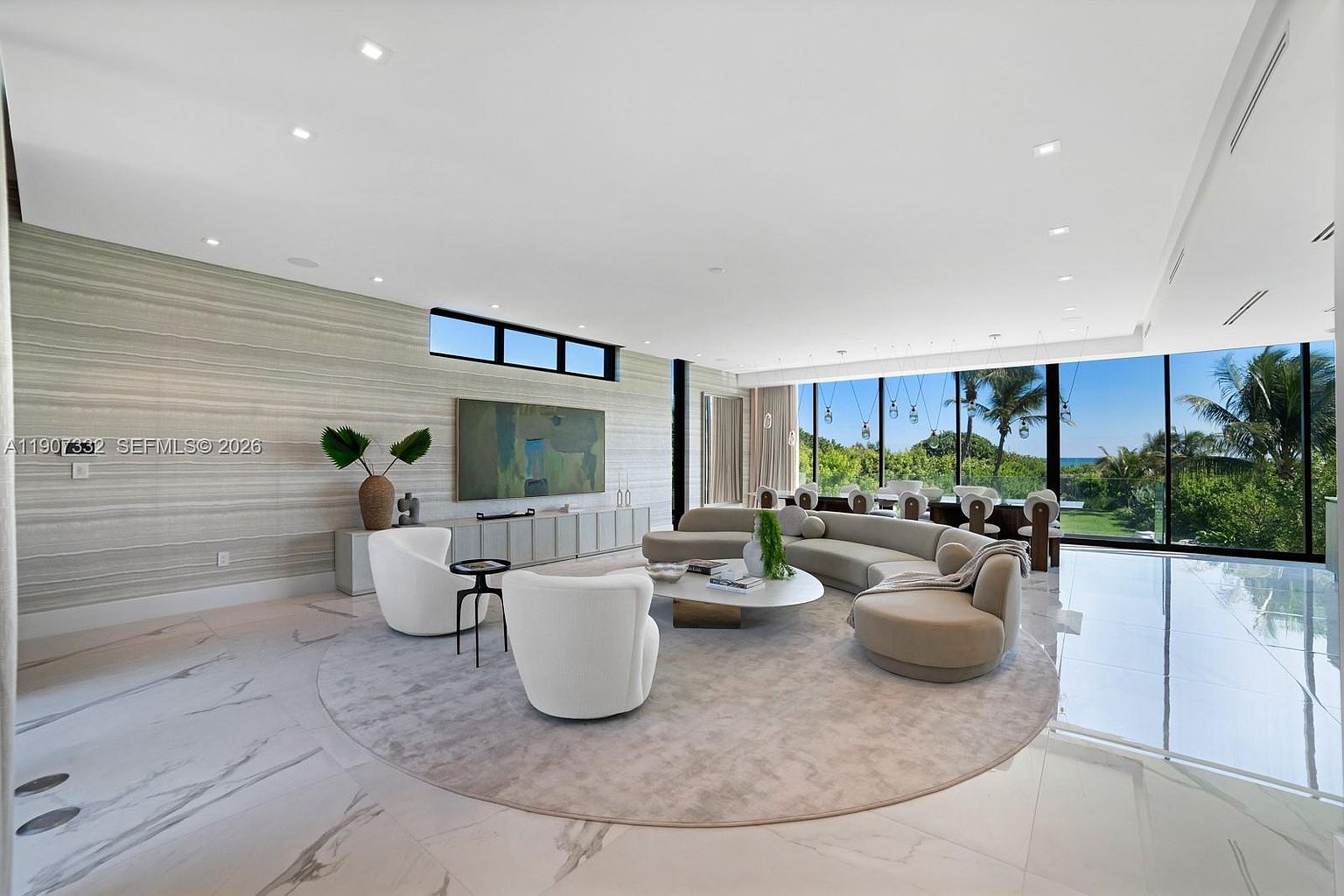 This expansive, open-concept living room features a sophisticated neutral palette with a large curved sectional sofa and two white accent chairs arranged on a circular area rug. The space is defined by a textured feature wall, a sleek media console, and floor-to-ceiling glass walls that provide panoramic views of the lush greenery and ocean beyond. The polished light-toned flooring and recessed lighting contribute to an airy, high-end contemporary aesthetic.