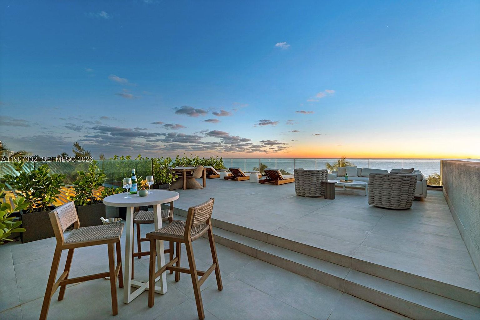 This expansive rooftop terrace offers a luxurious outdoor living space with panoramic ocean views at sunset. The area is furnished with a high-top dining set, multiple lounge chairs, and a comfortable seating arrangement, all set against a glass railing that maximizes the horizon line. The neutral-toned stone flooring and modern, woven furniture create a sophisticated and serene atmosphere perfect for entertaining.