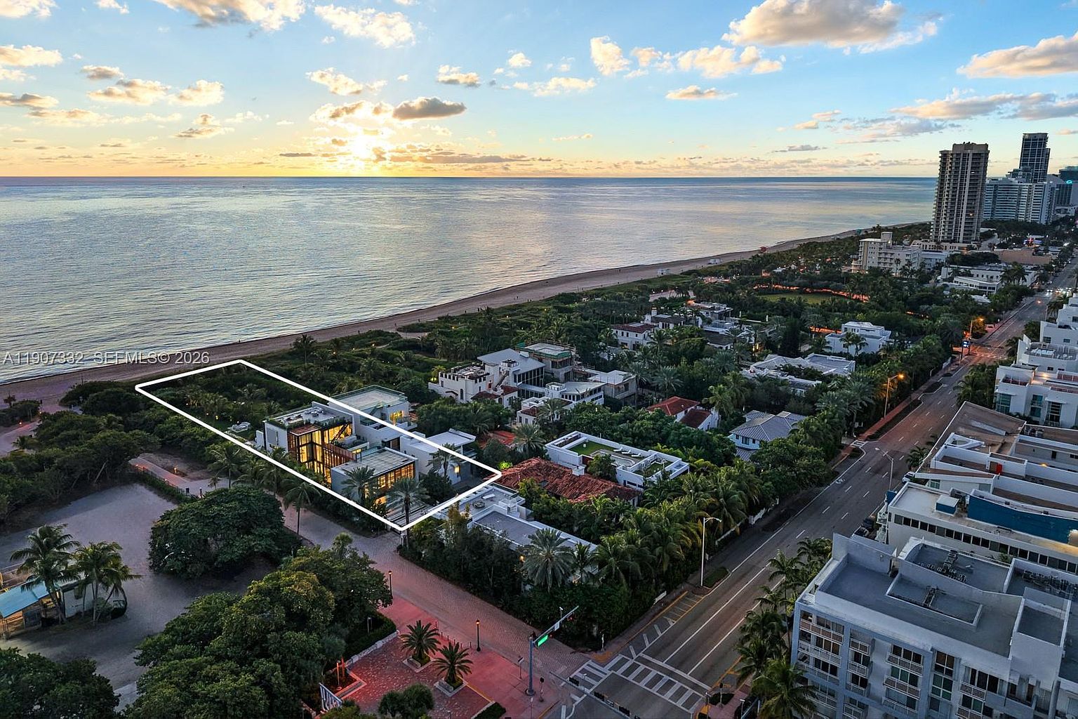 This high-angle aerial view captures a modern, multi-level residence situated in a prime coastal location, highlighted by a white digital outline. The property features a contemporary architectural design with flat roofs, expansive glass elements, and lush landscaping, positioned just steps from the sandy beach and ocean. The surrounding neighborhood includes dense tropical foliage, nearby high-rise buildings, and a coastal road, all bathed in the warm, golden light of a sunset.