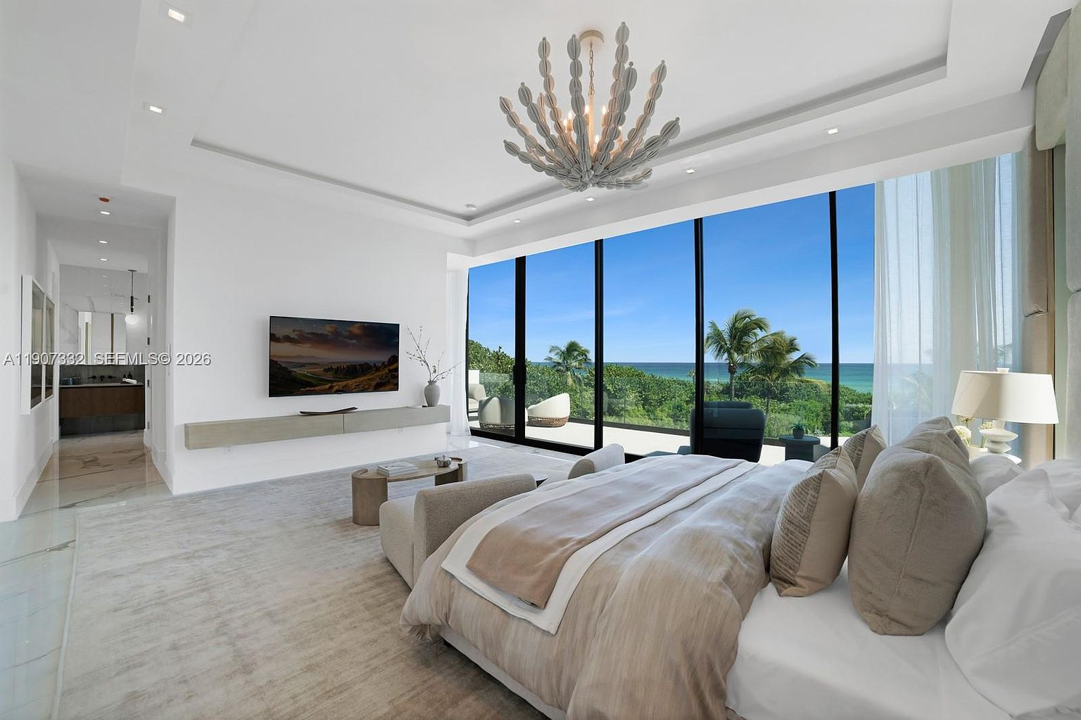 This spacious primary bedroom features a neutral color palette and a large bed centered in the room, facing a wall-mounted television and a floating media console. Floor-to-ceiling windows provide an expansive, unobstructed view of the ocean and lush greenery outside, while a unique, branch-like chandelier serves as a focal point on the ceiling. The room is bright and airy, with a minimalist aesthetic that emphasizes the connection to the coastal landscape.