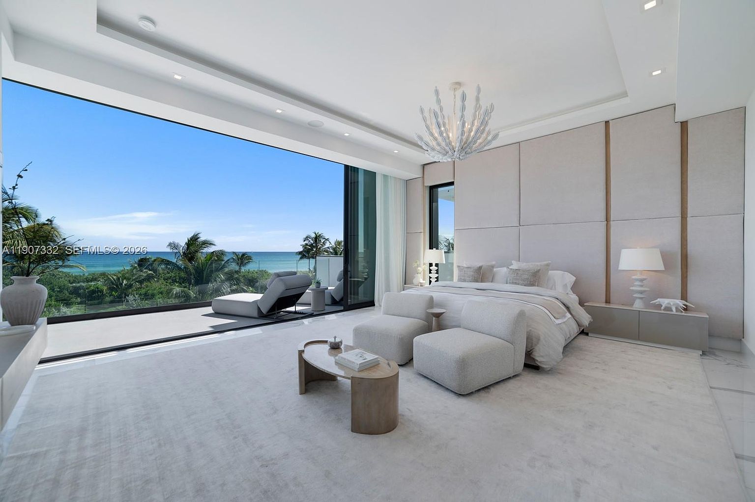 This expansive primary bedroom features a neutral, monochromatic palette and a minimalist aesthetic. The room is defined by floor-to-ceiling glass walls that provide an unobstructed, panoramic view of the ocean and balcony. A large bed with plush bedding is centered against a paneled wall, complemented by two modern armchairs and a low-profile coffee table, creating a serene and luxurious atmosphere.
