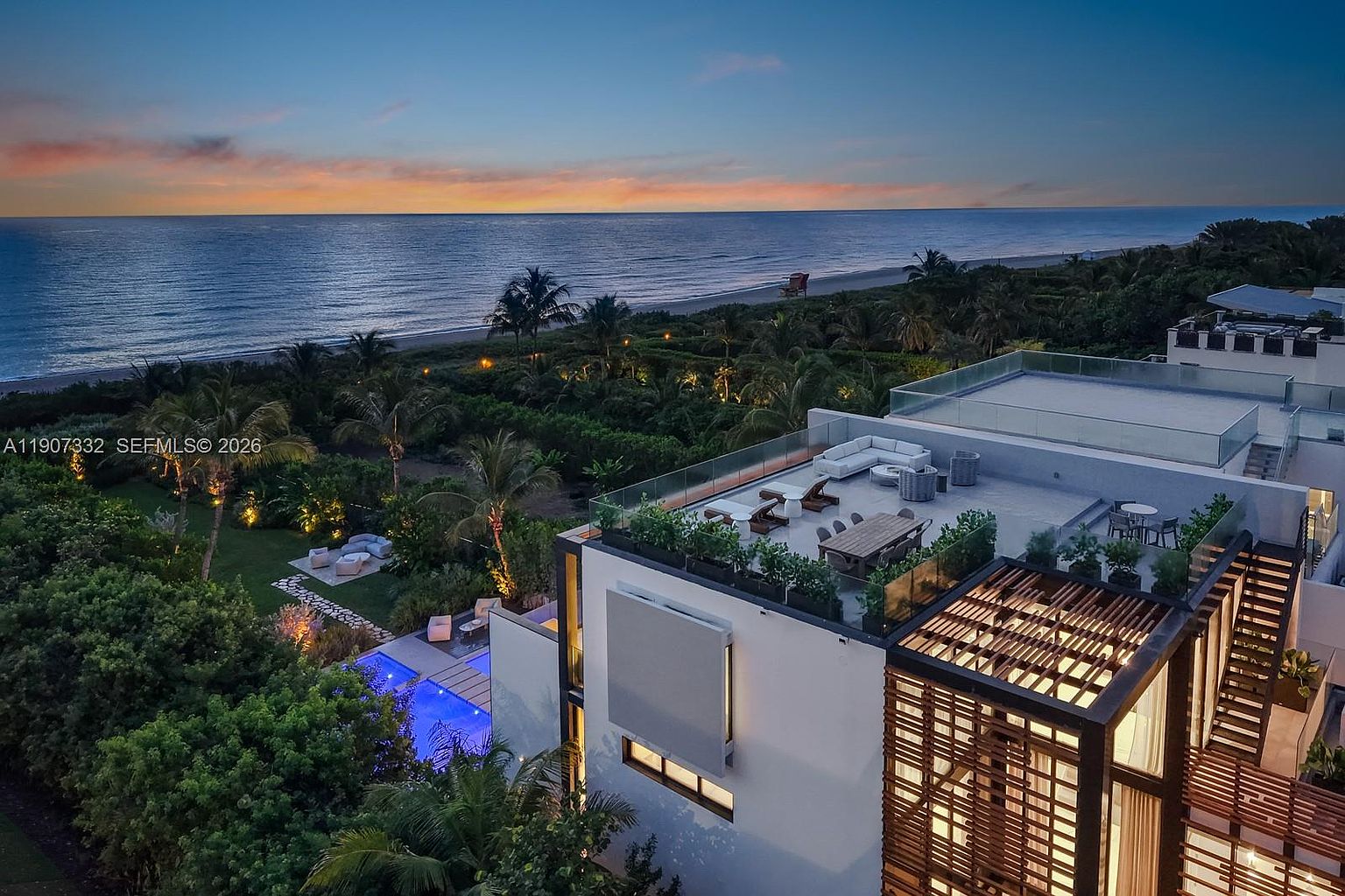 This high-angle aerial view captures a luxurious modern residence at dusk, situated directly along a serene coastline. The property features a spacious rooftop terrace furnished with lounge seating and a dining area, overlooking a private pool nestled within lush tropical landscaping. The scene is framed by the expansive ocean horizon and a vibrant sunset, emphasizing the home's prime waterfront location and sophisticated outdoor living spaces.