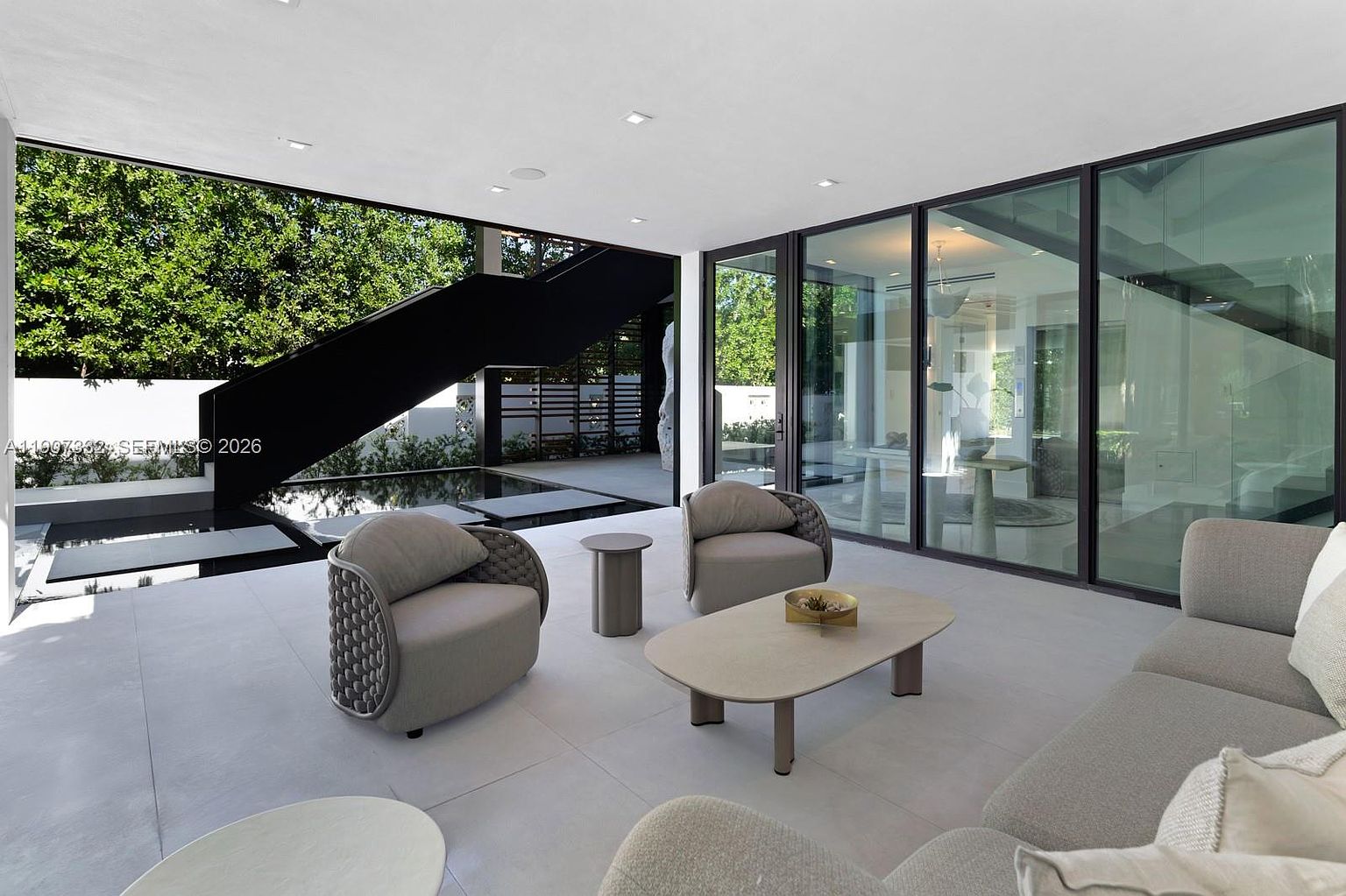 This modern, covered outdoor living space features a sleek, minimalist design with large glass walls that seamlessly connect the indoor and outdoor areas. The space is furnished with contemporary grey armchairs and a low-profile coffee table, overlooking a tranquil reflecting pool and a striking black staircase. The open-air layout creates a sophisticated, airy atmosphere perfect for high-end entertaining.