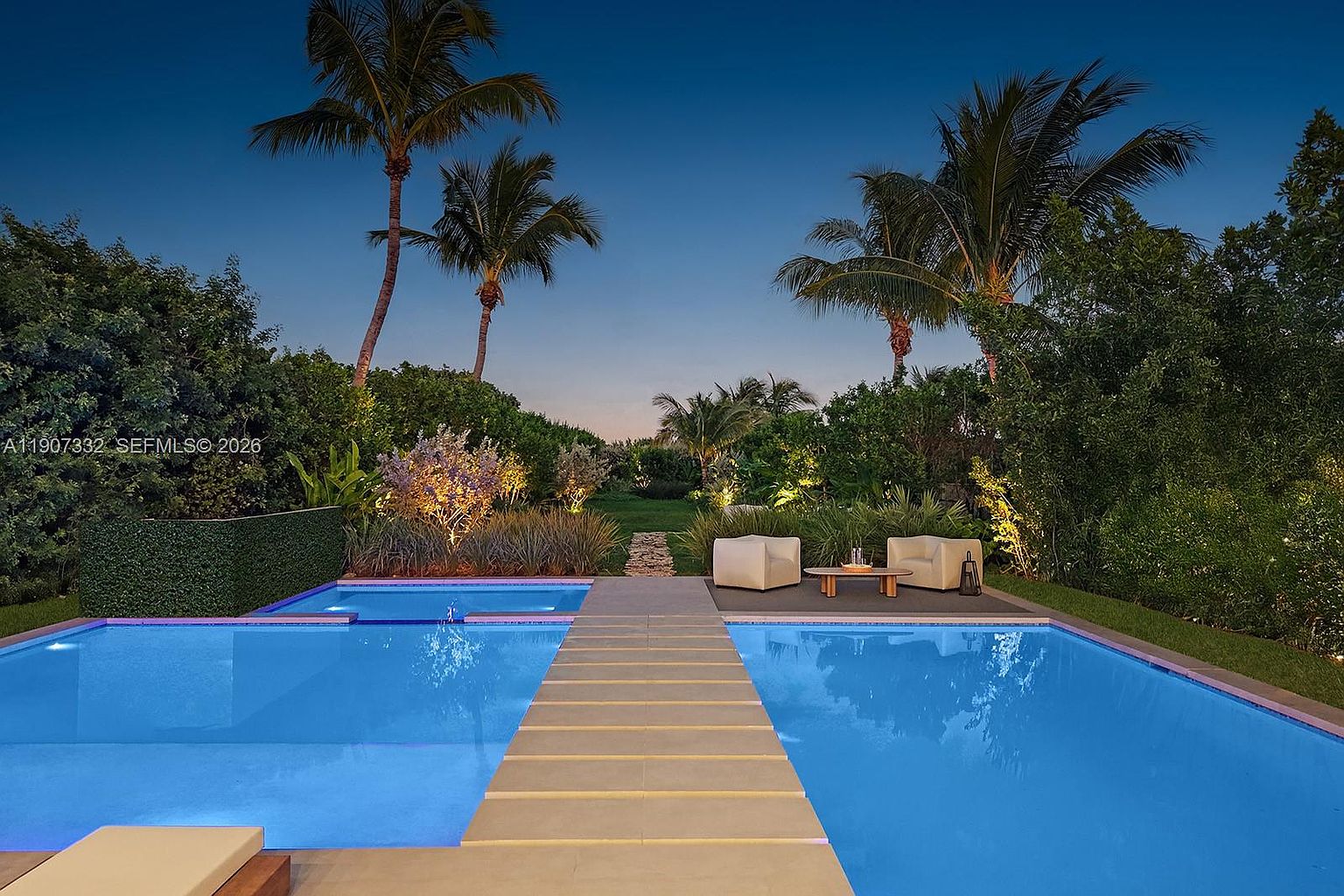 This serene backyard scene features a large, rectangular swimming pool with a central stone walkway bridge leading to a lush, landscaped garden. Two modern, light-colored lounge chairs sit on a deck area beside the pool, complemented by ambient landscape lighting that highlights the surrounding trees and foliage. The perspective is a low-angle, eye-level shot capturing the tranquil transition from the water to the private outdoor retreat at dusk.