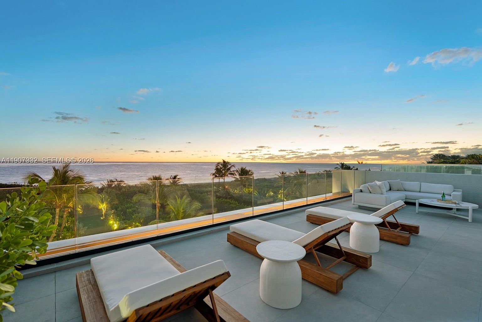 This expansive, modern balcony offers a serene outdoor living space with a panoramic view of the ocean at sunset. The area is furnished with sleek wooden lounge chairs, small side tables, and a comfortable sectional sofa, all set against a minimalist glass railing. The clean, neutral-toned flooring and open-air design create a sophisticated atmosphere perfect for relaxation and taking in the coastal scenery.