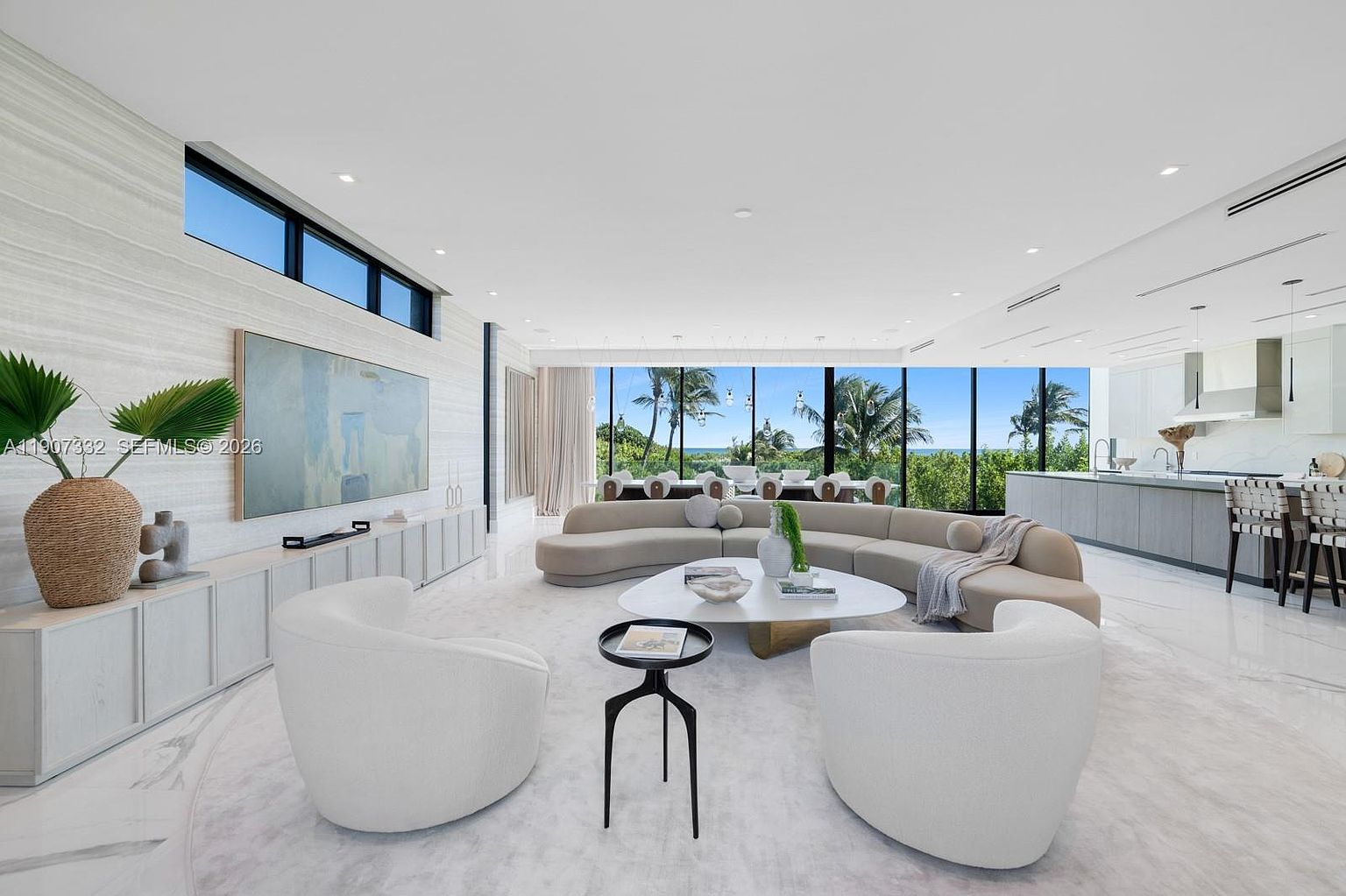This expansive, open-concept living area features a curved, neutral-toned sofa centered around a large, organic-shaped coffee table. The space is bathed in natural light from floor-to-ceiling windows that offer a panoramic view of the lush greenery and ocean beyond. A minimalist aesthetic is maintained with light-colored flooring, sleek cabinetry, and a large piece of abstract art, creating a sophisticated and airy atmosphere.