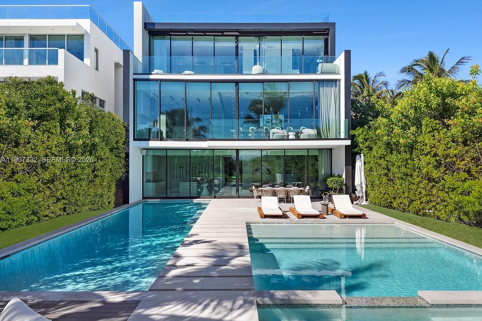 This modern, multi-story residence features a striking contemporary design with extensive glass walls that provide a seamless connection between the interior and the outdoor living space. The rear exterior showcases a long, narrow swimming pool and a separate lounge area with sun loungers on a stone deck, all framed by lush, mature privacy hedges. The clean lines and transparent facade create an airy, sophisticated aesthetic, emphasizing a luxurious indoor-outdoor lifestyle.