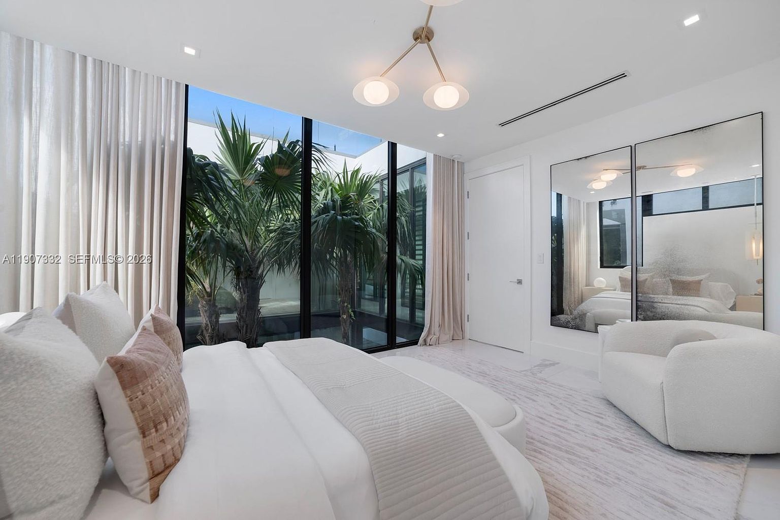 This modern primary bedroom features a minimalist aesthetic with a neutral color palette and plush white bedding. Large floor-to-ceiling windows provide a view into a private, lush courtyard filled with palm trees, while a large mirror reflects the room's serene atmosphere. A contemporary light fixture hangs from the ceiling, and a soft, textured rug anchors the space, creating an elegant and tranquil retreat.