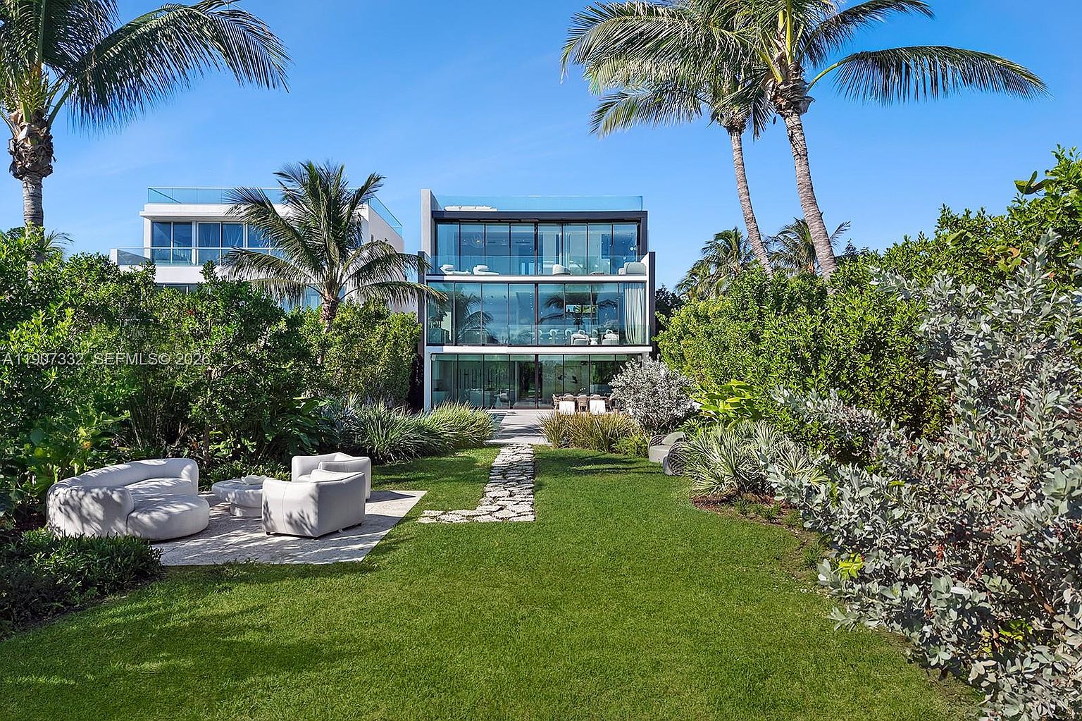 This modern, multi-story residence features a striking contemporary design with extensive glass walls that offer a seamless connection to the outdoors. The property includes a lush, manicured lawn with a stone pathway leading toward the house, complemented by comfortable outdoor lounge seating on a paved patio. Tall palm trees and dense, vibrant landscaping frame the structure, creating a private and serene tropical backyard oasis.