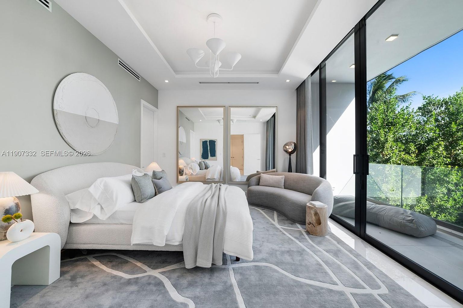 This sophisticated primary bedroom features a neutral color palette with soft textures, including a curved upholstered bed and a matching crescent-shaped sofa. The room is anchored by a large, patterned area rug and illuminated by a modern, sculptural chandelier suspended from a tray ceiling. Expansive floor-to-ceiling glass doors provide seamless access to a private balcony, flooding the space with natural light and offering views of lush greenery.