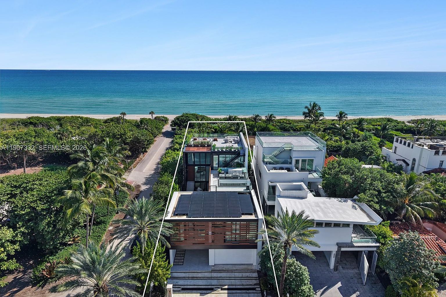 This high-angle aerial shot captures a modern, multi-level beachfront residence with a flat roof featuring solar panels and a rooftop terrace. The property is situated between lush tropical vegetation and a sandy beach, showcasing a contemporary architectural design with clean lines and large glass surfaces. The perspective highlights the home's prime location, its linear layout, and the seamless transition from the private residence to the coastal landscape.