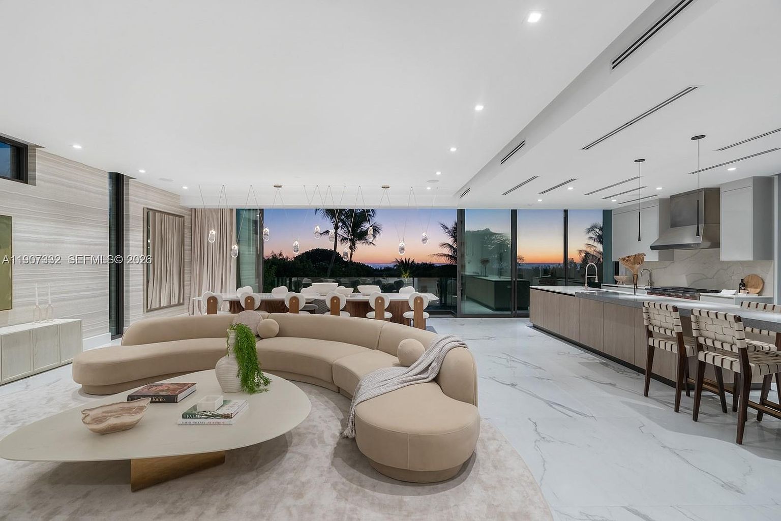 This expansive open-concept living area features a large, curved beige sofa centered on a textured rug, complemented by a low-profile coffee table. To the right, a sleek kitchen with light wood cabinetry and a stone-topped island provides a seamless transition for entertaining. The space is defined by floor-to-ceiling glass walls that frame a stunning sunset view over the horizon, creating a bright and sophisticated atmosphere.