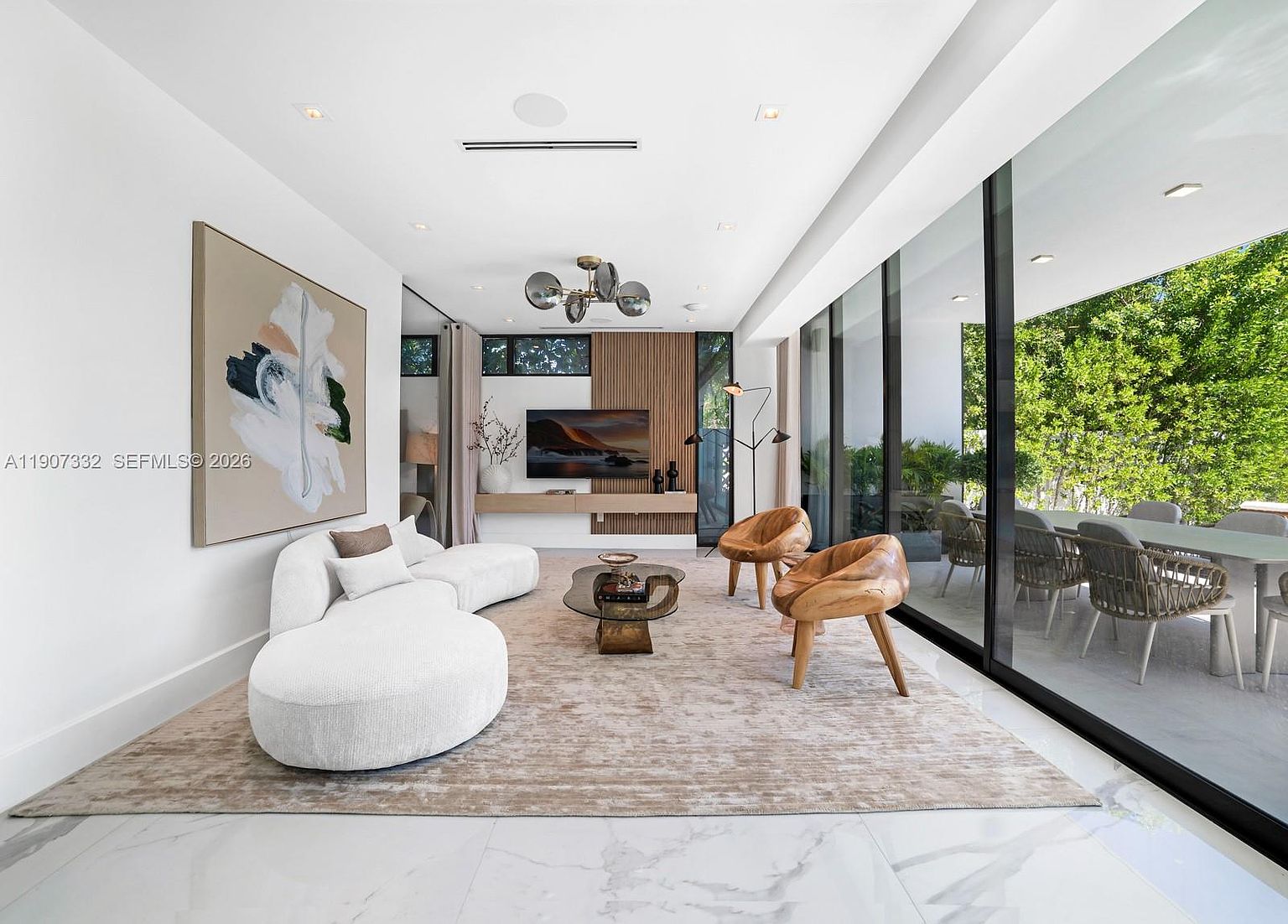 This modern living room features a curved white sofa and two sculptural wooden armchairs arranged on a textured area rug. A wall-mounted television is set against a vertical wood-slat accent wall, while floor-to-ceiling glass panels provide a seamless transition to an outdoor dining area. The space is finished with polished light-colored flooring and a contemporary multi-globe chandelier, creating an airy and sophisticated atmosphere.