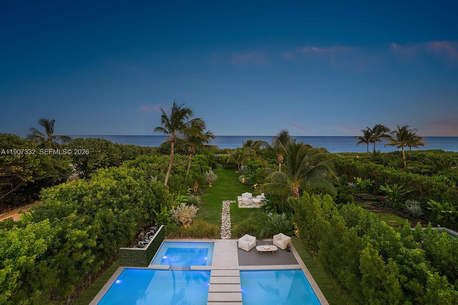 This high-angle aerial view captures a luxurious backyard oasis featuring a multi-section swimming pool with integrated lounge areas and a stone-paved path. Beyond the pool, a lush green lawn stretches toward a dense line of tropical trees and shrubs, which frame a direct, unobstructed view of the ocean horizon. The scene is set during the twilight hour, creating a serene and sophisticated atmosphere perfect for outdoor relaxation.