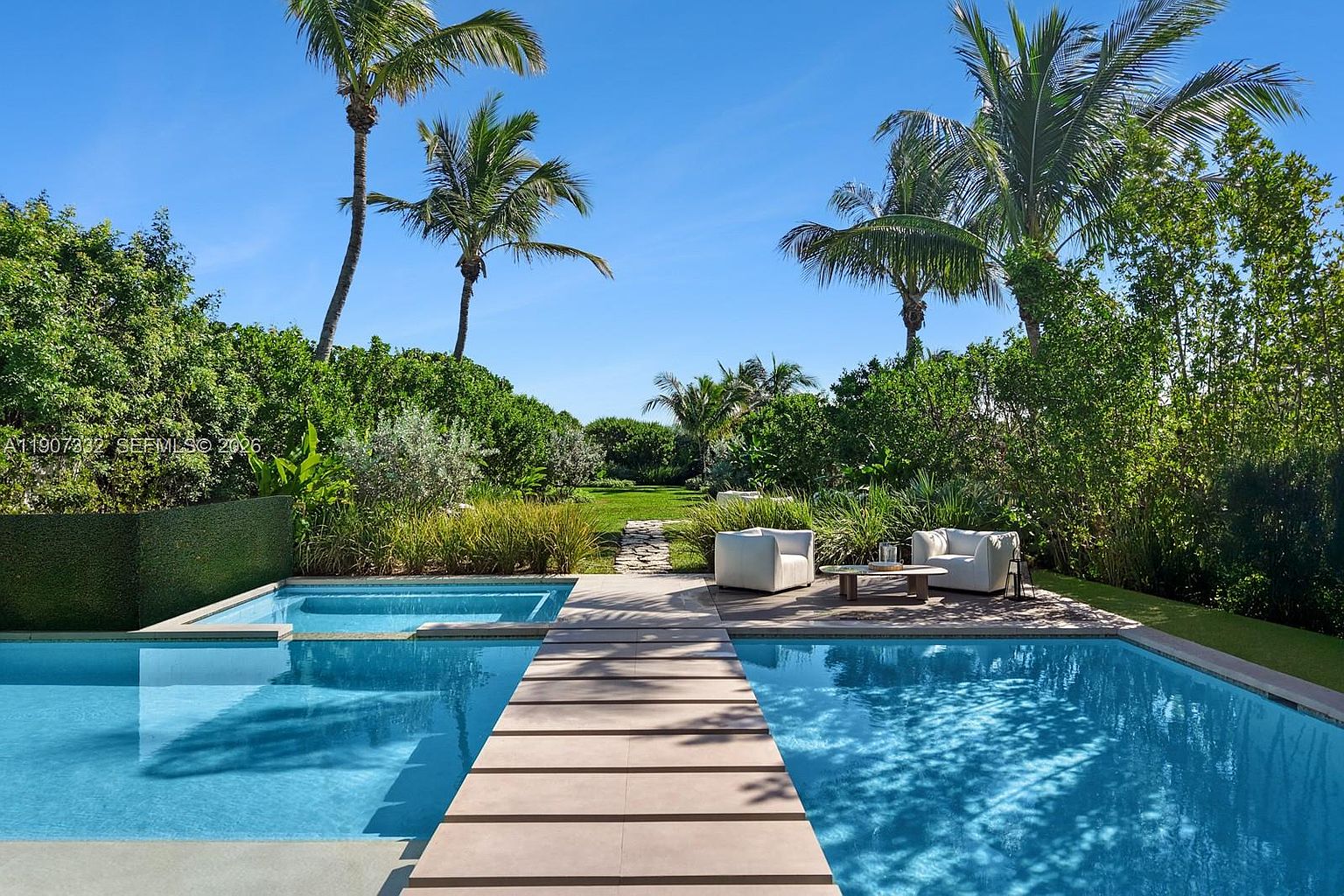 This serene outdoor space features a rectangular swimming pool with a central stone walkway bridge leading toward a lush, manicured lawn. To the right, a modern seating area with two white armchairs and a low coffee table sits on a wooden deck, surrounded by dense tropical greenery and tall palm trees. The bright blue sky and clear water create a tranquil, resort-like atmosphere perfect for relaxation.