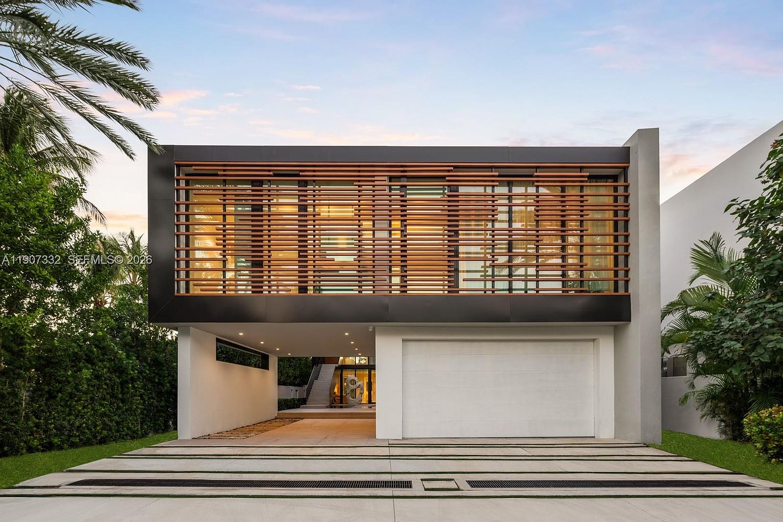 This modern, two-story residence features a striking cantilevered upper level adorned with horizontal wood slats that provide privacy and texture. The ground floor includes a white garage door and an open-air covered entryway leading to a staircase, set against a backdrop of lush tropical landscaping. The paved driveway features integrated linear drainage and lighting, contributing to the home's clean, contemporary architectural aesthetic.