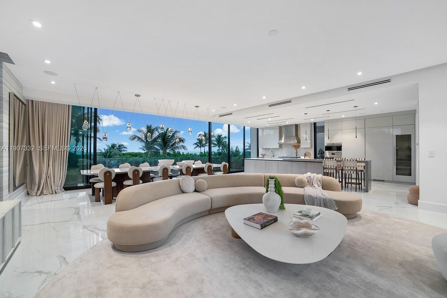 This expansive, open-concept living area features a large, curved beige sofa centered around a unique, organic-shaped coffee table. The space is defined by polished light-toned flooring and floor-to-ceiling windows that provide a panoramic view of the lush palm trees and sky outside. A dining area and a sleek, modern kitchen with a central island are seamlessly integrated into the same open floor plan, creating a bright and airy atmosphere.