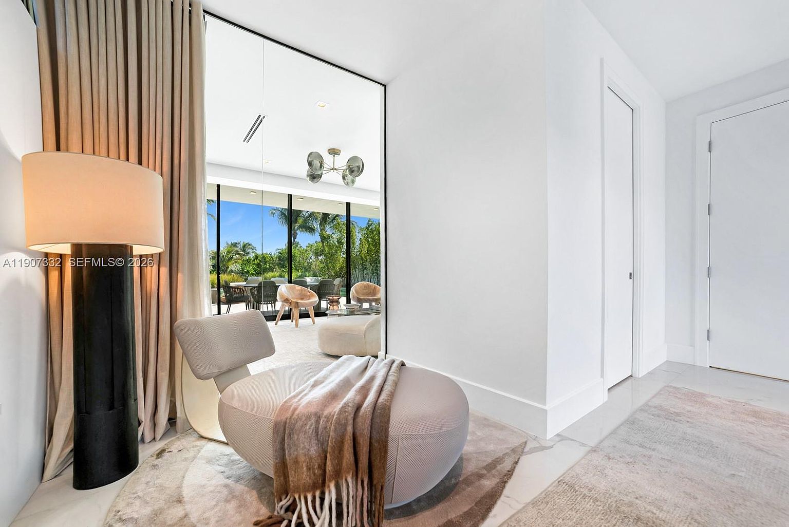 This bright, modern living space features a minimalist lounge chair draped with a soft throw blanket, positioned on a textured area rug. A tall, dark floor lamp stands beside floor-to-ceiling curtains, while a large, framed mirror reflects an outdoor patio area with dining furniture and lush greenery. The room is characterized by clean white walls and a polished light-colored floor, creating an airy and sophisticated atmosphere.