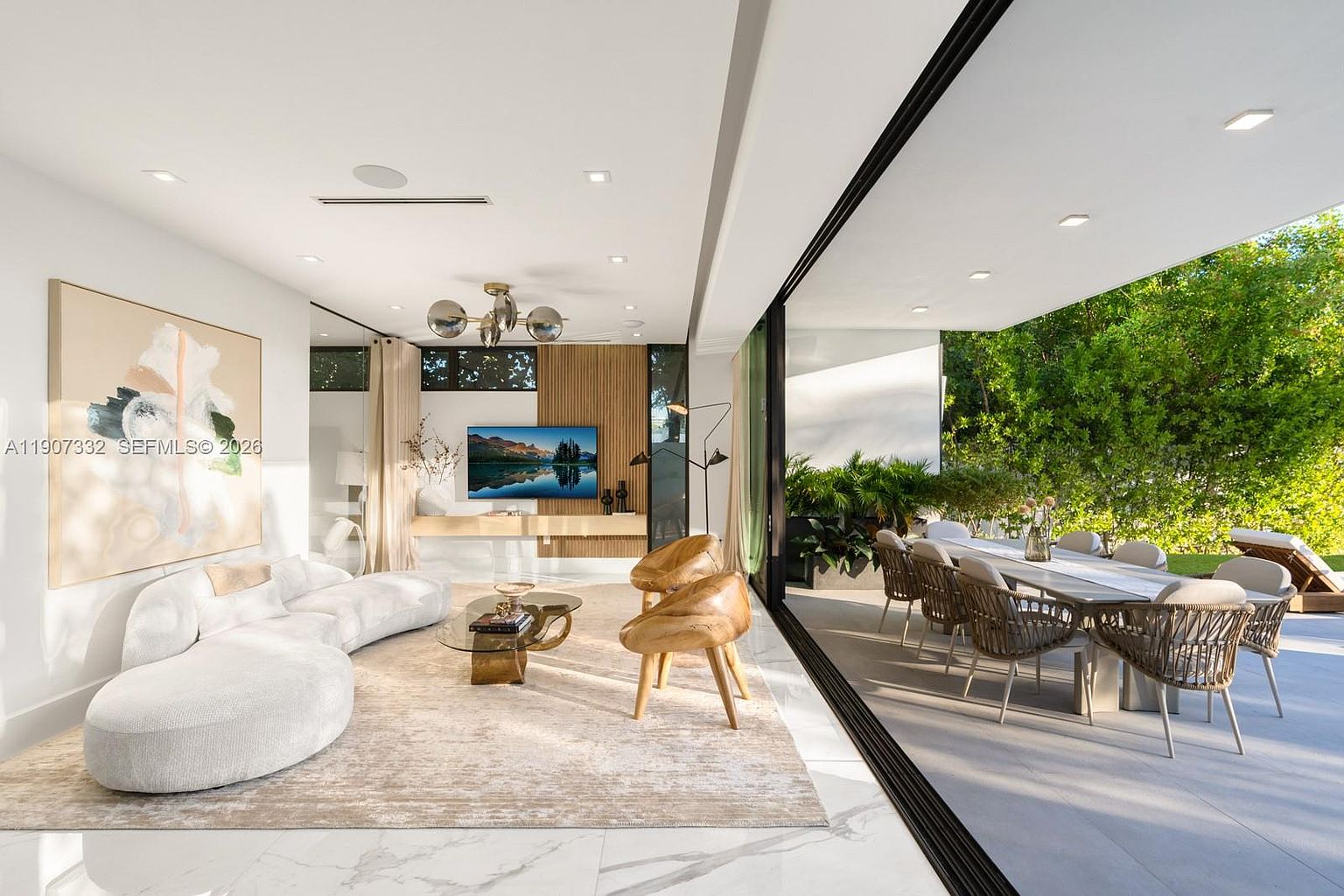 This bright, open-concept living room features a curved white sofa, two sculptural wooden accent chairs, and a glass-topped coffee table set on a textured area rug. The space seamlessly transitions to an outdoor dining area through expansive floor-to-ceiling glass sliding doors, creating a fluid indoor-outdoor living experience. A large abstract painting adorns the wall, while a television is mounted on a light wood-paneled feature wall, contributing to the sophisticated and airy aesthetic.