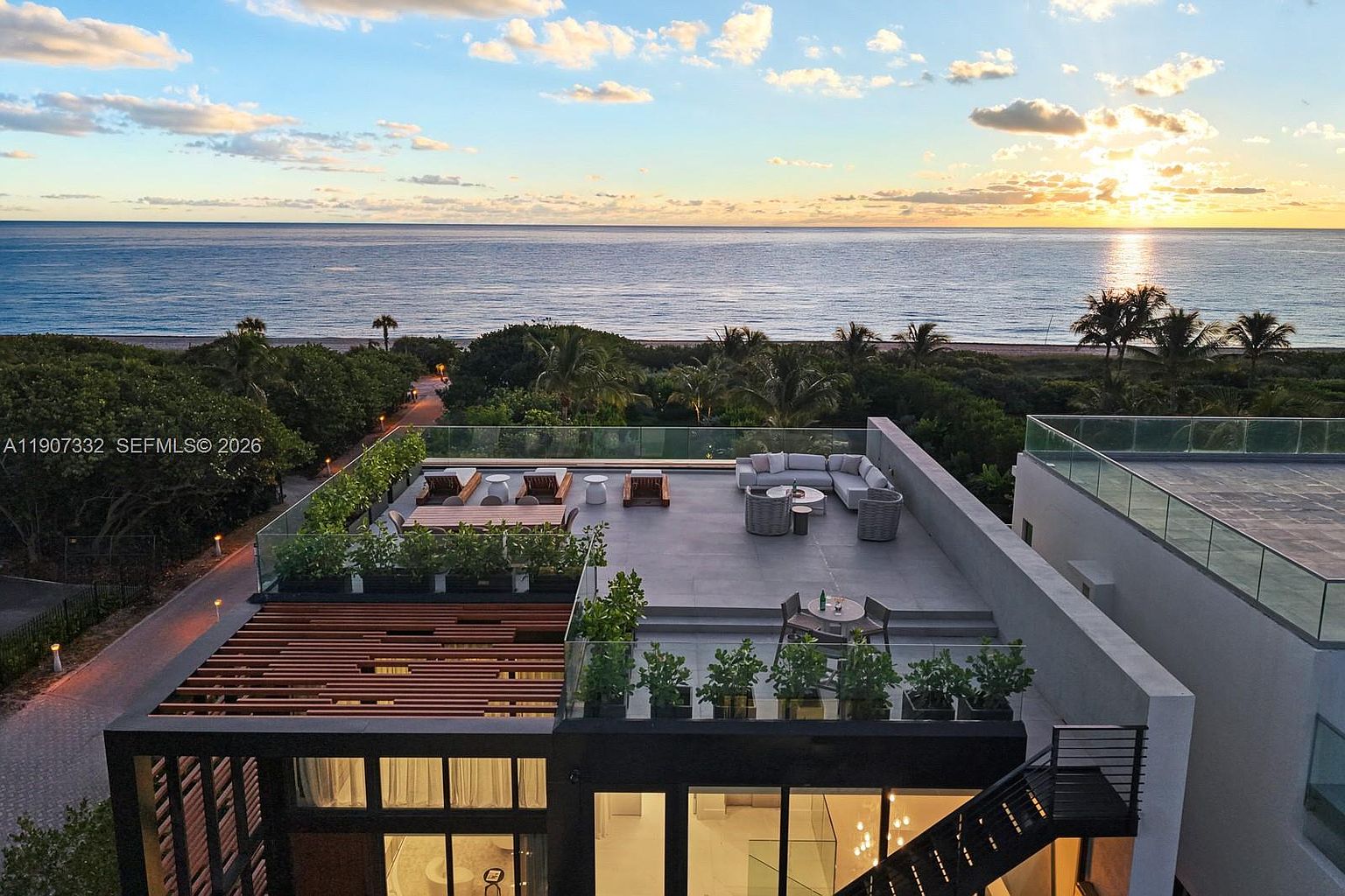 This high-angle aerial view captures a modern rooftop terrace overlooking the ocean at sunset. The expansive deck features a comfortable lounge area with modular seating, a dining space, and a wooden pergola, all framed by glass railings and lush greenery. The composition emphasizes the seamless transition between the luxury outdoor living space and the serene coastal horizon.