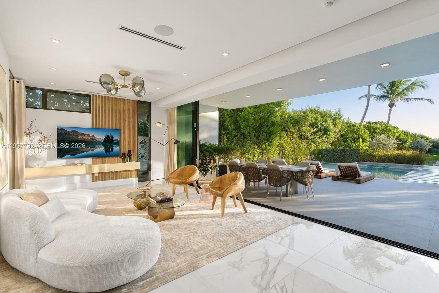 This expansive, modern living room features a curved white sofa, two wooden accent chairs, and a glass-topped coffee table set upon a textured area rug. The space seamlessly transitions to an outdoor patio and pool area through large, retractable glass walls that provide an unobstructed view of the lush greenery and palm trees. A wall with vertical wood paneling serves as a focal point for the television, while a unique, multi-globe chandelier adds a sculptural element to the high ceiling.