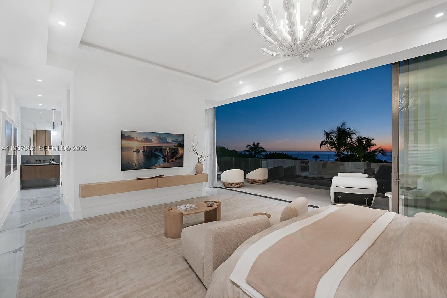This luxurious primary bedroom features a neutral color palette and a seamless transition to an expansive outdoor balcony through large sliding glass doors. The room is anchored by a plush bed and a minimalist light wood floating console beneath a wall-mounted television, while a unique, sculptural chandelier serves as a focal point above. The space offers a serene atmosphere with a direct, unobstructed view of the ocean at dusk, complemented by polished light-toned flooring.