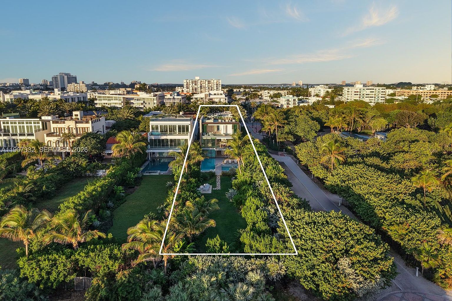 This high-angle aerial view captures a modern, multi-story residence nestled within a lush, tropical landscape. The property features a private swimming pool, a spacious green lawn, and a white outline highlighting the expansive lot boundaries. The surrounding area is densely wooded, providing a sense of privacy while maintaining proximity to the nearby urban skyline.