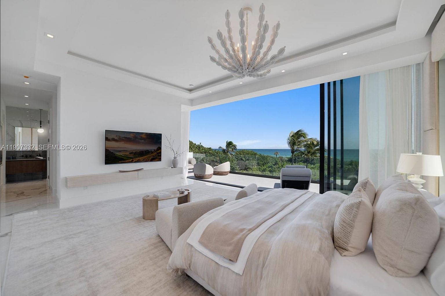 This luxurious primary bedroom features a neutral, monochromatic color palette and a seamless transition to an expansive outdoor balcony through large, floor-to-ceiling glass sliding doors. The room is anchored by a plush bed with layered linens, a unique sculptural chandelier, and a minimalist media console mounted below a wall-mounted television. The open-concept design emphasizes the panoramic ocean views, creating a serene and sophisticated atmosphere.