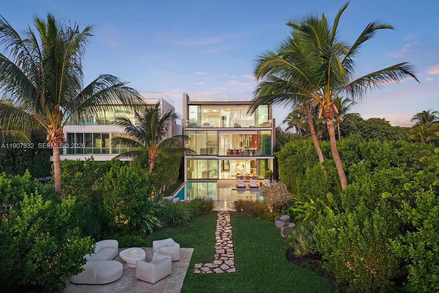 This modern three-story residence features a sleek, contemporary design with extensive glass walls that offer a seamless connection between the interior and the outdoor living space. The property includes a private swimming pool, a stone-paved patio area, and a lush, manicured lawn framed by mature palm trees and dense tropical landscaping. The perspective is captured from an elevated position, showcasing the home's architectural symmetry and its serene, resort-like backyard setting at dusk.