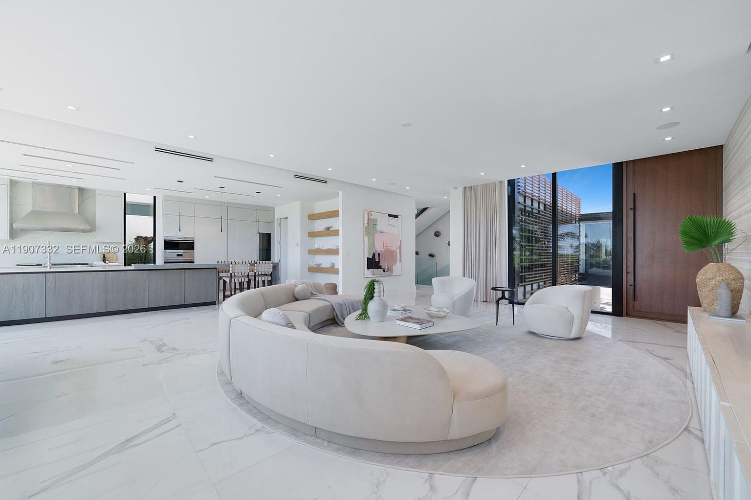 This expansive, open-concept living area features a large, curved sectional sofa arranged on a circular area rug, creating a sophisticated focal point. The space is characterized by bright, neutral tones, polished stone flooring, and a seamless transition to the outdoors through large glass panels. A minimalist kitchen with a long island and bar seating is visible in the background, emphasizing the home's airy and modern architectural design.