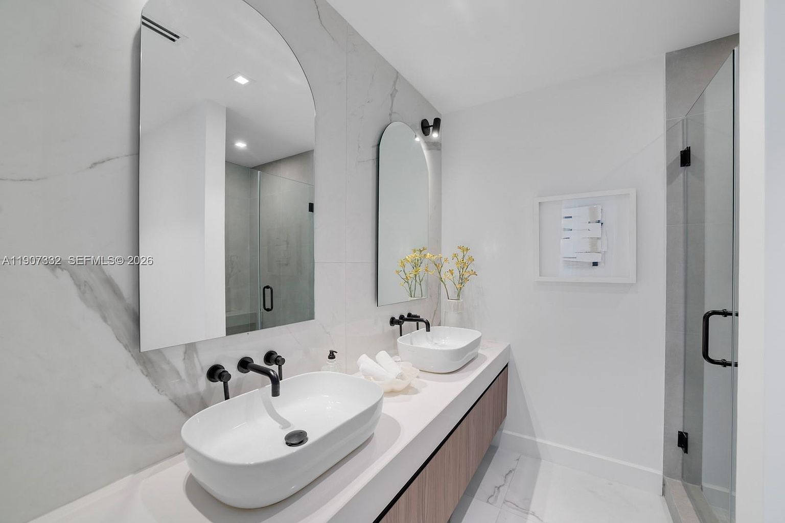 This modern primary bathroom features a double vanity with two vessel sinks and matte black fixtures set against a large, light-veined stone backsplash. Two arched mirrors are mounted above the vanity, complemented by minimalist wall lighting and a floating wood-toned cabinet. To the right, a glass-enclosed shower adds to the sleek, contemporary aesthetic of the space.