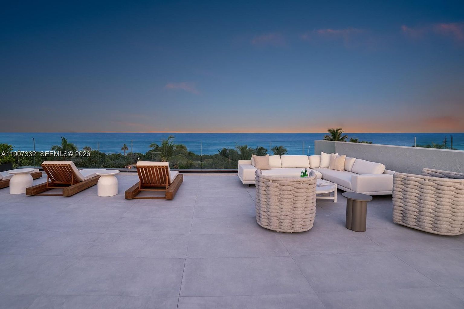 This expansive rooftop terrace offers a luxurious outdoor living space with panoramic ocean views at dusk. The area is furnished with a large white sectional sofa, woven armchairs, and wooden lounge chairs, all set against a backdrop of the sea and a clear sky. A glass railing provides an unobstructed view, creating a seamless connection between the terrace and the horizon.