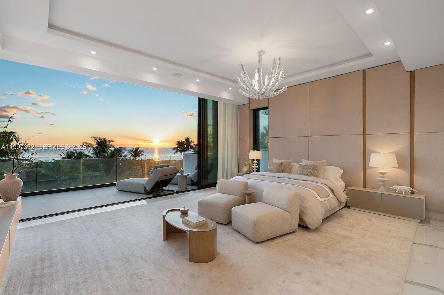 This luxurious primary bedroom features a neutral, monochromatic palette with soft textures and a large, upholstered wall panel behind the bed. Floor-to-ceiling glass doors open onto a private balcony, framing a stunning sunset view over the ocean and palm trees. The room is furnished with a plush bed, two modern armchairs, and a sleek coffee table, all illuminated by a striking, sculptural chandelier.