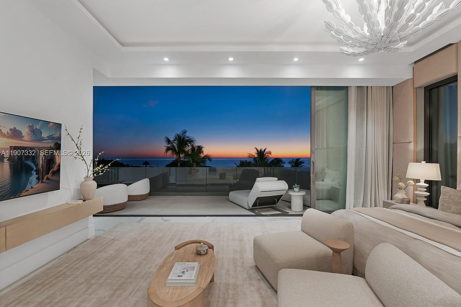 This luxurious primary bedroom features a neutral color palette and a seamless transition to a private balcony through expansive floor-to-ceiling glass doors. The room is furnished with plush, contemporary seating and a low-profile bed, all oriented toward the stunning sunset view over the ocean. A unique, sculptural chandelier hangs above, adding a sophisticated touch to the serene and airy atmosphere.