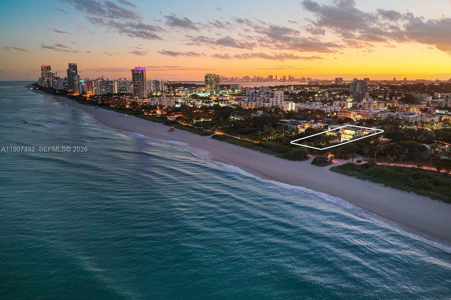 This high-altitude aerial shot captures a luxury beachfront property at sunset, highlighted by a white outline for emphasis. The perspective showcases the home's prime location directly on the coastline, with the vast ocean on one side and a sprawling urban skyline in the distance. The warm, golden hour lighting enhances the serene atmosphere of the coastal landscape.