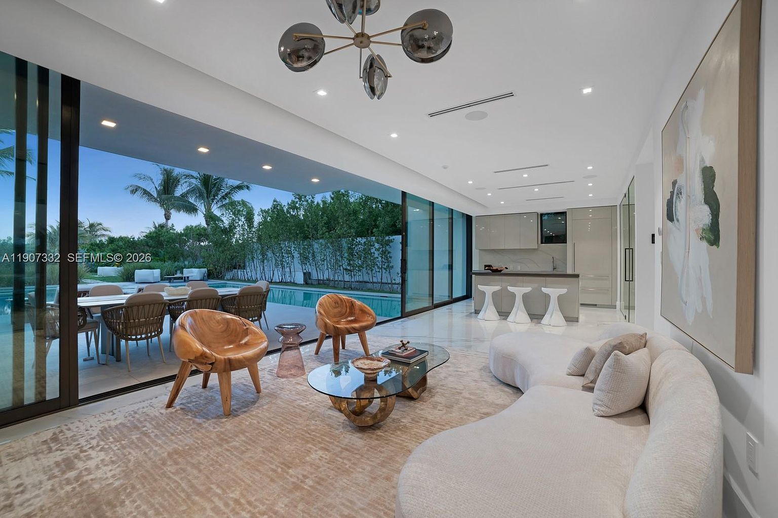 This modern open-concept living area features a curved, light-colored sofa and two sculptural wooden armchairs arranged around a glass-topped coffee table. The space seamlessly transitions to an outdoor patio and pool area through expansive floor-to-ceiling sliding glass doors. In the background, a sleek kitchen with a breakfast bar and white stools completes the bright, airy, and sophisticated interior design.