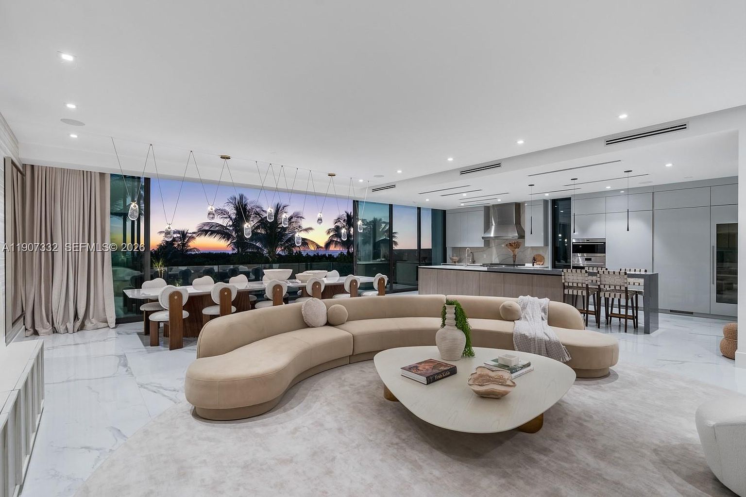 This expansive, open-concept living area features a large, curved beige sofa centered around a unique triangular coffee table on a plush area rug. The space is defined by sleek, light-toned flooring and a seamless transition to a dining area and kitchen, all illuminated by recessed lighting and a series of modern pendant lights. Large glass walls provide an unobstructed view of a sunset over the ocean, creating a sophisticated and airy atmosphere.