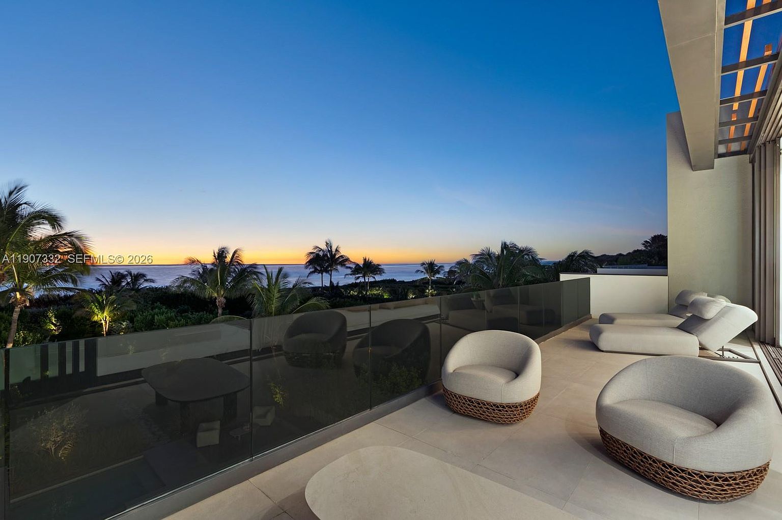 This expansive, modern balcony offers a serene outdoor living space with a clear glass railing that provides unobstructed views of the ocean at sunset. The area is furnished with comfortable, rounded lounge chairs and a low-profile table, creating a sophisticated atmosphere for relaxation. The scene is captured from a slightly elevated perspective, emphasizing the seamless transition between the indoor living area and the tranquil coastal horizon.