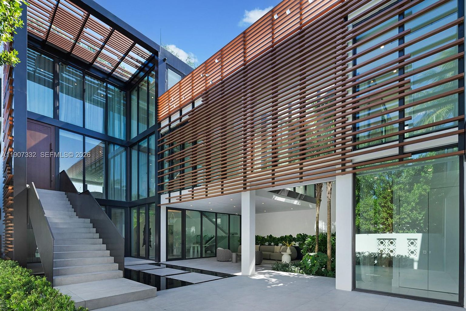 This modern architectural residence features a striking combination of dark metal framing, expansive glass walls, and horizontal wood slat screens that provide both texture and privacy. A concrete staircase leads to an elevated entrance, while a covered outdoor lounge area sits beneath the upper level, overlooking a serene water feature and lush greenery. The design emphasizes clean lines, indoor-outdoor flow, and a sophisticated, contemporary aesthetic.