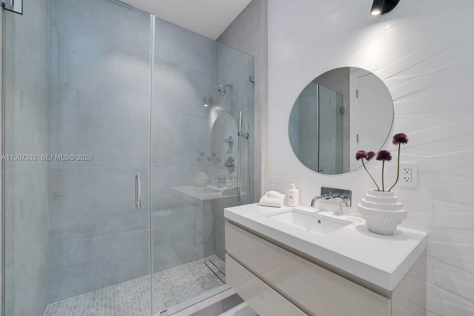 This modern bathroom features a sleek, wall-mounted vanity with a white countertop and a circular mirror centered on a textured white wall. To the left, a spacious walk-in shower is enclosed by glass panels and finished with light-toned wall tiles and a mosaic-tiled floor. The space is accented by a decorative vase with purple flowers, creating a clean, contemporary, and minimalist aesthetic.