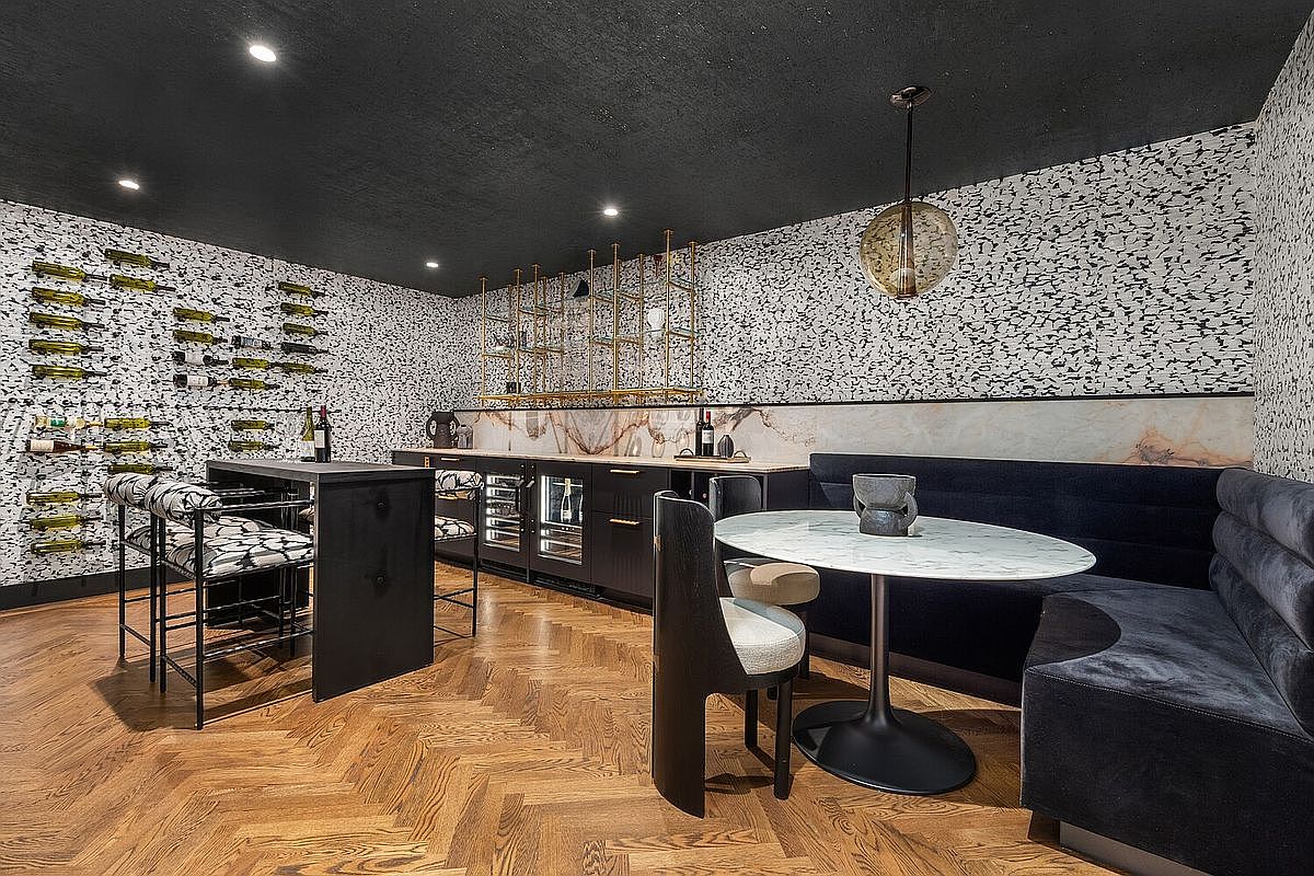This interior shot showcases a stylish wine cellar featuring a unique wine rack wall display, a bar area with built-in wine coolers, and a cozy seating nook with a round table. The room is decorated with patterned wallpaper and a modern pendant light, creating an inviting and sophisticated atmosphere perfect for entertaining.