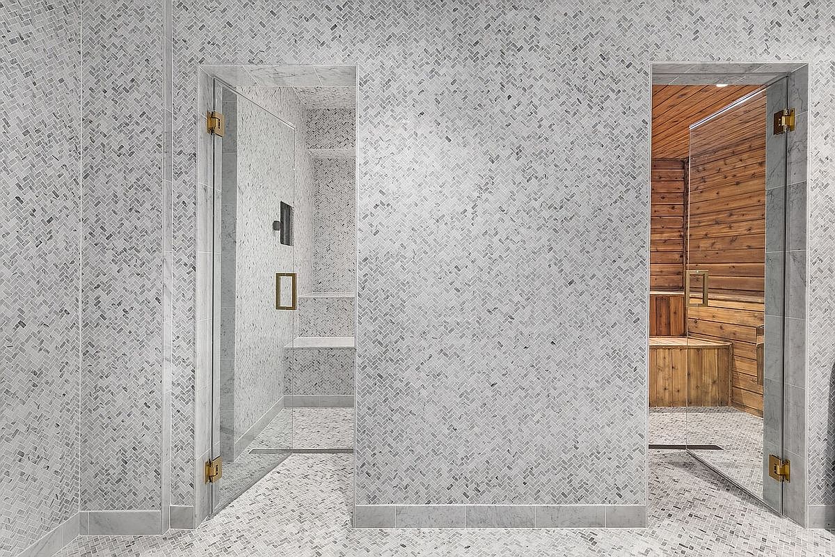 This image showcases a luxurious primary bathroom featuring mosaic tile walls and flooring. Two glass-enclosed spaces are visible: a shower on the left and a sauna on the right, both with gold hardware. The overall impression is one of high-end spa-like comfort and modern elegance.